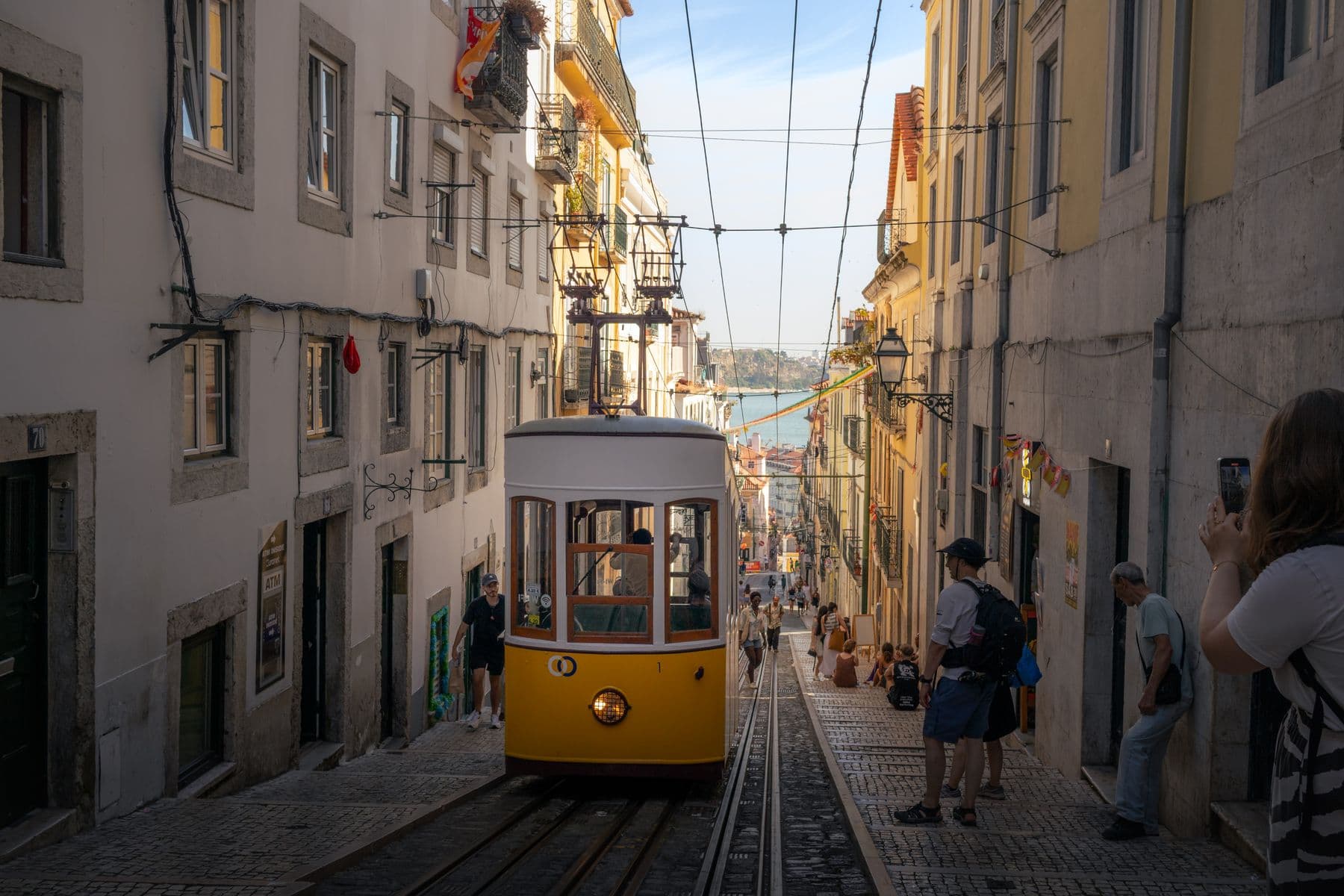 A yellow Lisbon tram on a steep street leading towards the Tagus